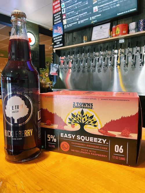A bottle of huckleberry cider next to a box of raspberry lemonade cider cans on a wooden bar top.