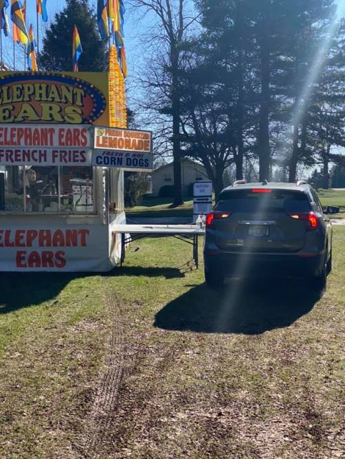 A car parked near a food stand selling elephant ears, French fries, lemonade, and corn dogs on a sunny day.