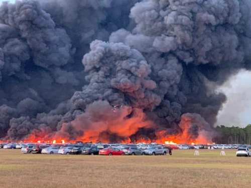 A large plume of black smoke rises over a field with parked cars, indicating a significant fire in the background.
