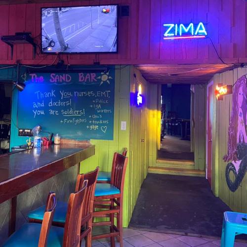 A colorful bar interior with a neon "ZIMA" sign, a TV on the wall, and a message thanking first responders.