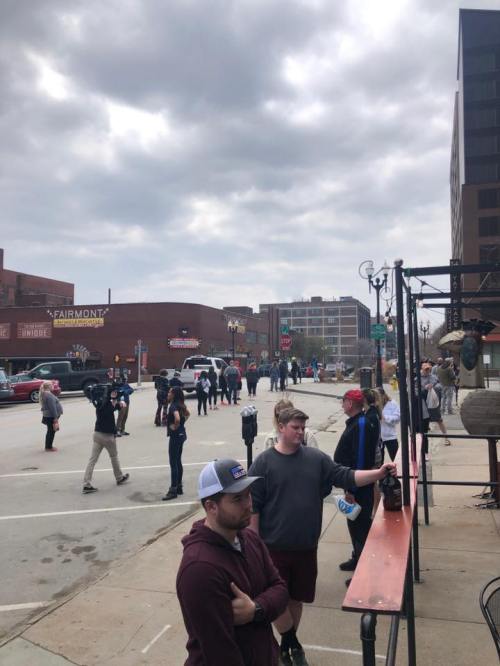 A crowd gathers on a city street with buildings in the background under a cloudy sky. Some people are standing near a bar.