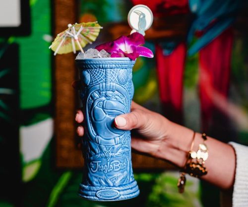 A hand holds a blue tiki mug filled with a colorful cocktail, adorned with an umbrella and a flower.