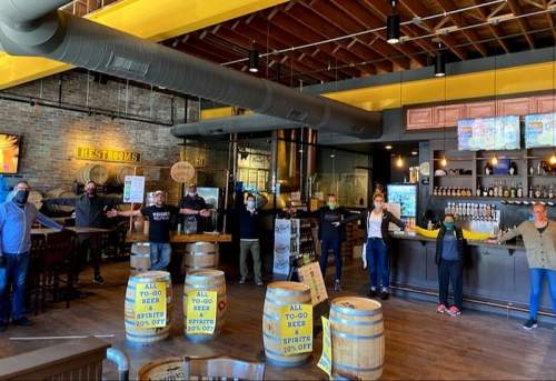 A group of people in a bar, standing with arms outstretched, promoting to-go beer and spirits.