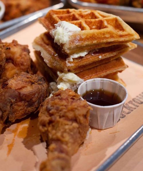 A stack of waffles with butter and syrup, accompanied by crispy fried chicken on a tray.