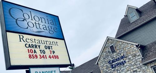 Sign for Colonial Cottage restaurant, featuring carry-out hours and contact number, with a stone building in the background.
