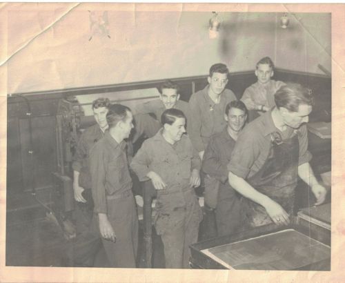 A group of men in work uniforms gathered in a vintage printing shop, smiling and interacting with each other.