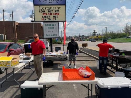 Three people in masks and gloves prepare food outside a restaurant under a sign, with tables and cooking equipment nearby.