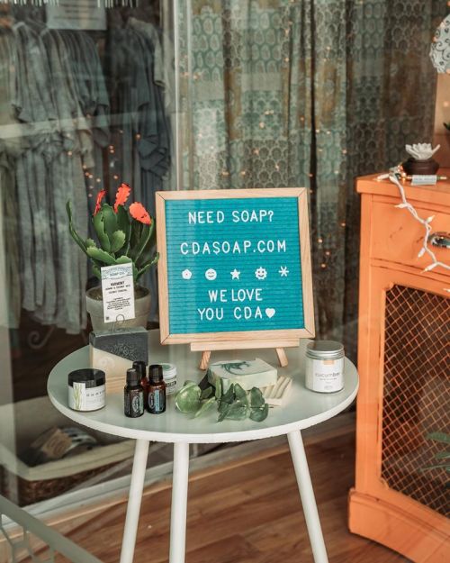 A small table displays a sign for CDA Soap, with plants and products, in a cozy shop setting.