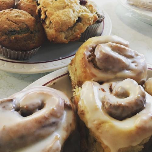 A plate of cinnamon rolls with icing and a tray of muffins in the background.