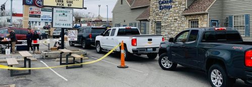 Cars lined up for curbside pickup outside a restaurant, with tables and a sign indicating the service.