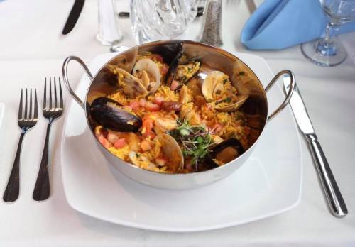 A silver bowl filled with seafood paella, featuring shrimp, mussels, and clams, on a white tablecloth with utensils.