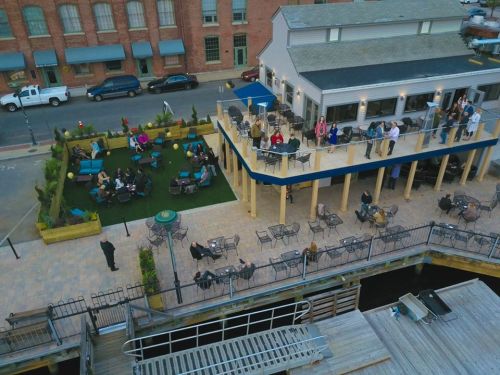 Aerial view of a lively outdoor dining area with people socializing on a deck and patio by the water.
