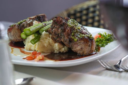 A plate of grilled meat with asparagus, mashed potatoes, and a drizzle of sauce, garnished with tomatoes and greens.