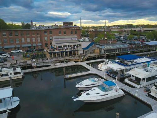 Aerial view of a marina with boats docked, surrounded by buildings and a cloudy sky at sunset.