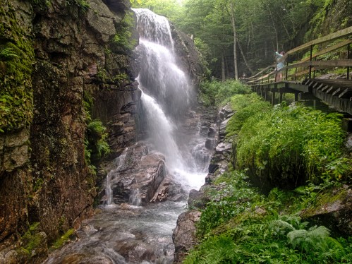 A serene waterfall cascades down rocky cliffs, surrounded by lush greenery and a wooden walkway.