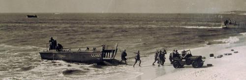 Soldiers disembark from a landing craft onto a beach, with a jeep and boats in the background. Historical wartime scene.