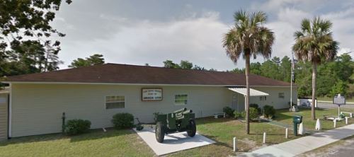 A single-story building with a sign reading "American Legion," featuring a cannon in front and palm trees nearby.