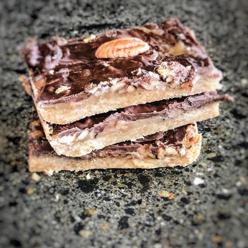 Three layers of chocolate-covered dessert bars with a pecan on top, resting on a dark speckled surface.