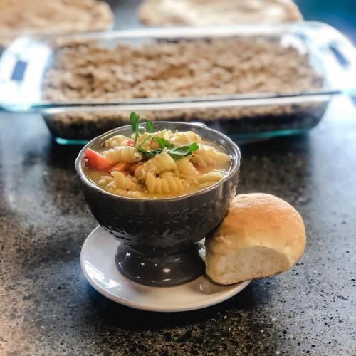 A bowl of pasta soup with vegetables, served with a roll, and a casserole dish in the background.