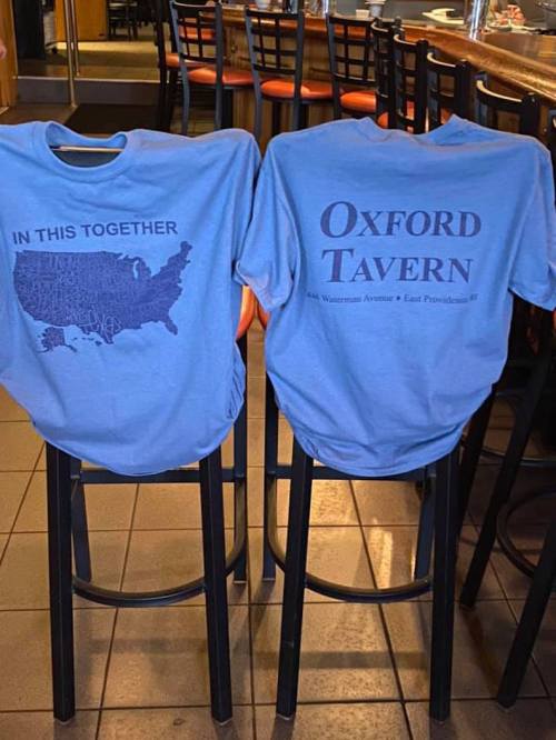 Two blue t-shirts hanging on bar stools, one with a map of the USA and the text "IN THIS TOGETHER," the other reading "Oxford Tavern."