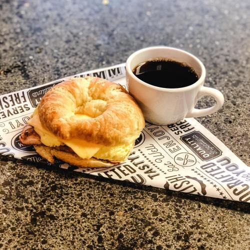A croissant sandwich with egg and cheese beside a cup of black coffee on a patterned paper.