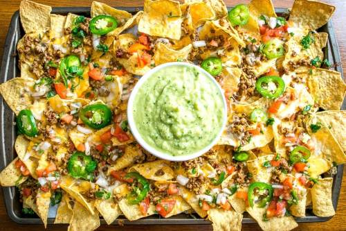 A large tray of nachos topped with cheese, ground beef, tomatoes, jalapeños, and a bowl of guacamole in the center.