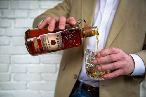 A person pours whiskey from a bottle into a glass, set against a white brick wall.