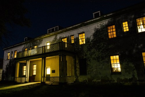 An old, weathered building at night, illuminated by warm light from its windows against a starry sky.