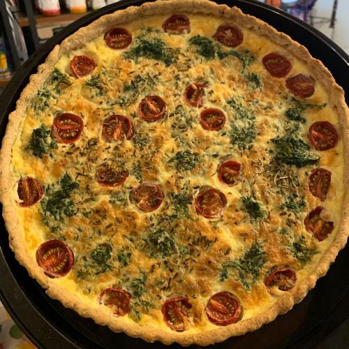 A freshly baked quiche with cherry tomatoes and herbs on a golden crust, placed on a black baking tray.