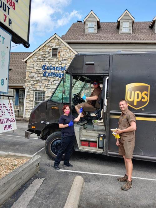 Two UPS drivers in uniforms and masks interact outside a delivery truck, with a building in the background.