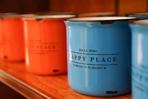 Colorful enamel mugs in orange and blue, labeled "Happy Place" with coordinates, displayed on a wooden shelf.