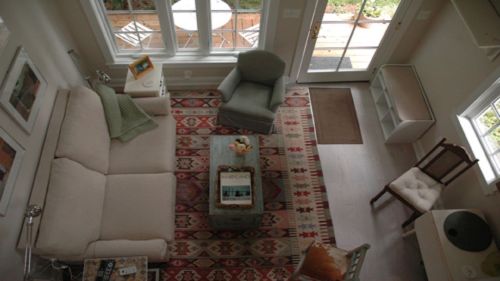 A cozy living room with a light sofa, patterned rug, coffee table, and large windows letting in natural light.