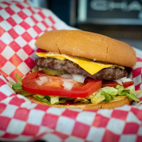 A close-up of a cheeseburger with lettuce, tomato, pickles, and onions in a checkered paper wrapper.