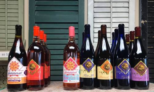 Bottles of wine with colorful labels displayed on a table against a backdrop of wooden shutters.