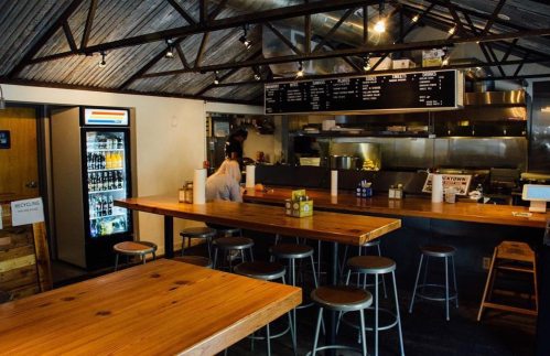 Interior of a casual eatery with wooden tables, metal stools, and a visible kitchen area. A person sits at the counter.