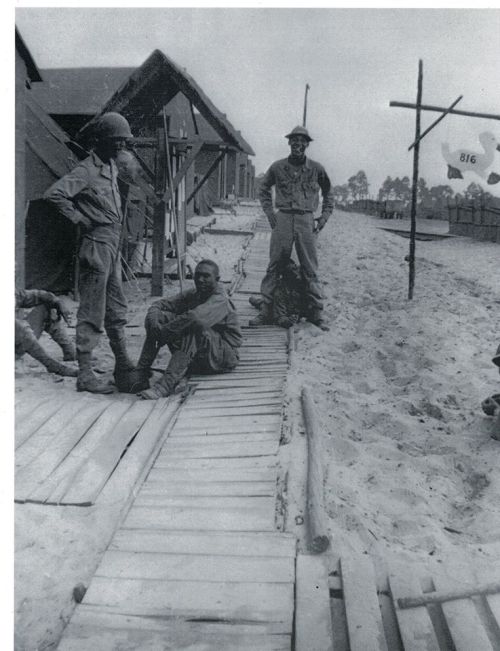 Black and white photo of soldiers on a sandy path near wooden structures, with one soldier sitting and two standing.