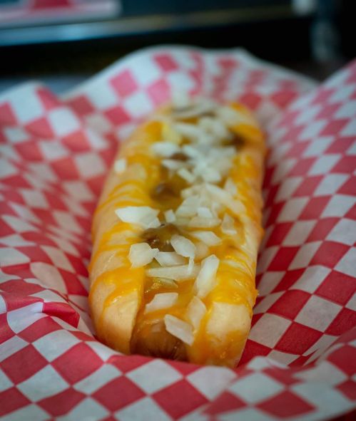 A hot dog topped with melted cheese and chopped onions, served in a red and white checkered paper basket.