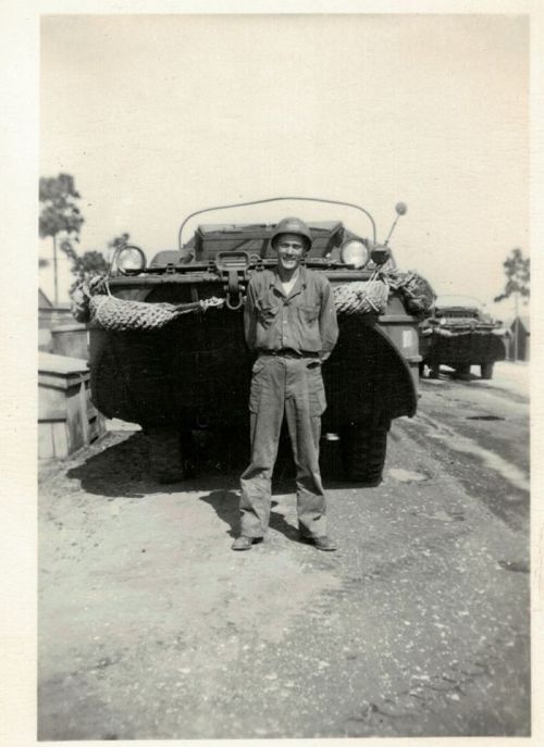 A soldier stands in front of a military vehicle, wearing a helmet and uniform, on a dirt road.