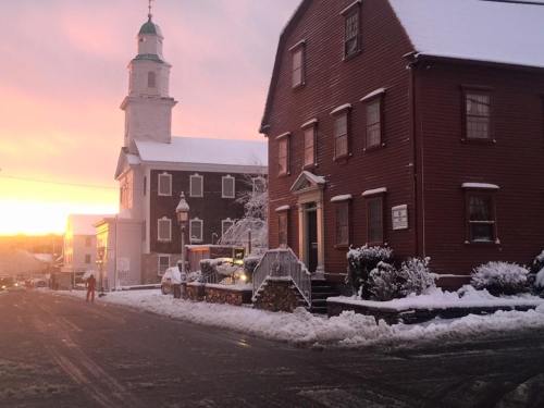 A snowy street scene at sunset, featuring a red house and a church with a green steeple.