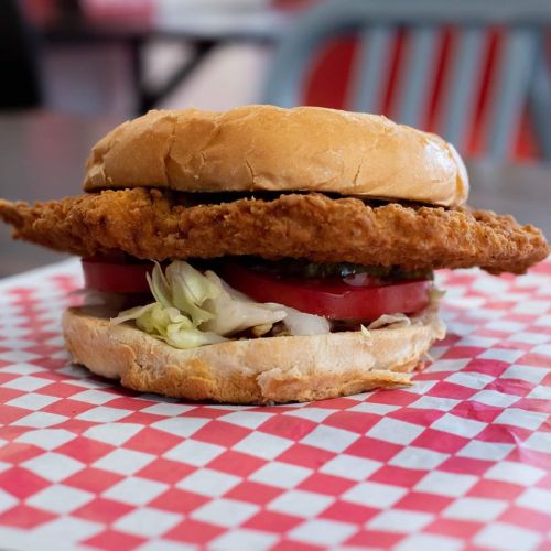 A crispy fried chicken sandwich with lettuce, tomato, and pickles on a toasted bun, served on a red-checkered paper.