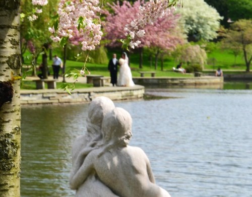 A couple in wedding attire by a serene lake, framed by cherry blossoms and a stone sculpture of two figures.