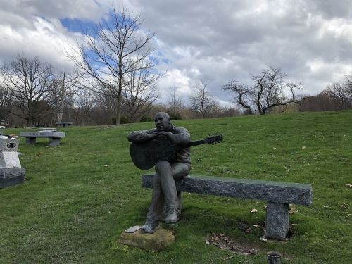 A bronze statue of a seated musician playing guitar on a stone bench in a grassy park under a cloudy sky.