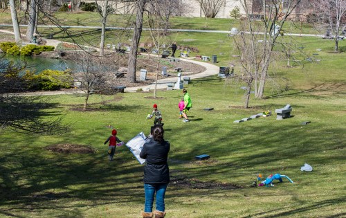 A woman stands on a hill overlooking children playing in a park with trees and a pond in the background.