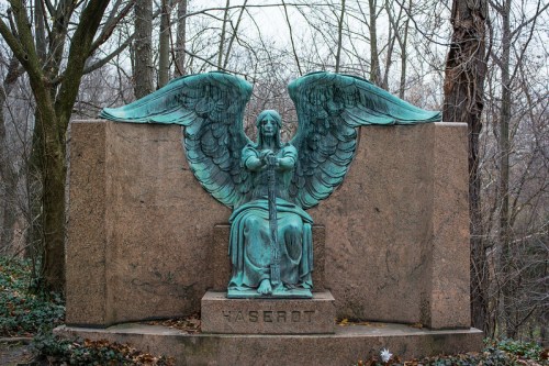 A bronze angel statue with wings, seated and holding a sword, surrounded by bare trees and a stone backdrop.