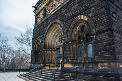 A stone building with intricate arches and carvings, featuring a staircase and bare trees in the background.