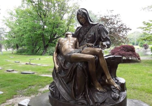 A bronze statue of a seated Mary holding the lifeless body of Jesus, set in a serene cemetery landscape.