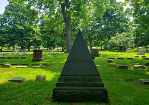 A pyramid-shaped gravestone in a lush green cemetery, surrounded by other gravestones and trees.
