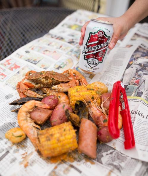 A hand holds a can of Narragansett beer above a spread of seafood, corn, and potatoes on newspaper.