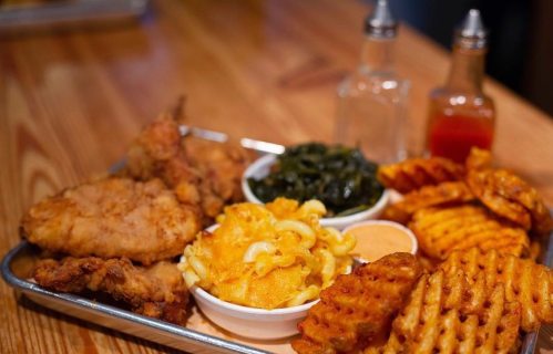 A metal tray with fried chicken, macaroni and cheese, collard greens, and waffle fries, with condiments in the background.