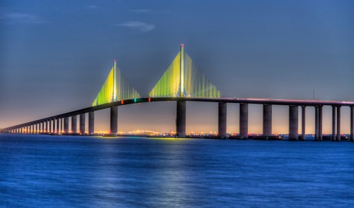 A brightly lit suspension bridge spans over calm waters at dusk, with colorful lights illuminating its towers.